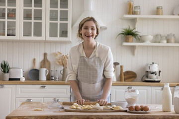 Happy beautiful young baker woman in apron preparing bakery food for order, baking at home, rolling dough on floury table with fresh ingredients, looking at camera, smiling. Female portrait