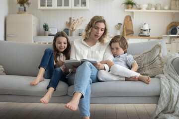 Young daycare teacher woman reading book to kids on home sofa. Caring mother telling fairytale to children on couch, teaching son and daughter to read, enjoying motherhood, educational activity