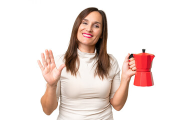 Middle-aged caucasian woman holding coffee pot over isolated background saluting with hand with happy expression