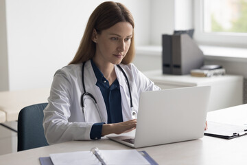 Positive focused pretty doctor woman typing on laptop at work table in clinic office. Physician,...