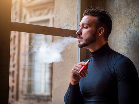 Handsome man vaping, smoking e-cigarette by a window