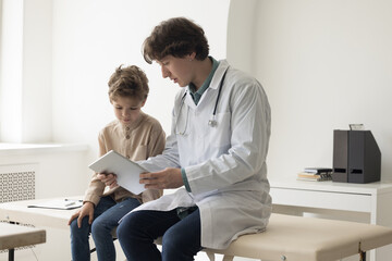 Engaged young pediatrician man showing content on tablet to patient kid, explaining examination result, diagnosis. Male doctor giving device to boy for playing, reading ebook.