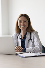 Happy pretty doctor woman sitting at work table in hospital, clinic office, looking at camera with toothy smile. Portrait of positive cheerful female practitioner, physician in white coat, stethoscope