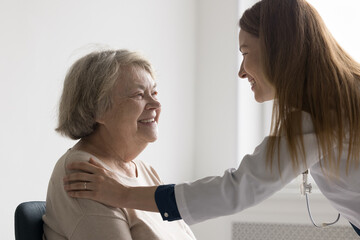 Fototapeta premium Happy pretty doctor woman giving comfort to positive older senior patient, touching shoulder, speaking, smiling, laughing. Elder lady visiting practitioner in office