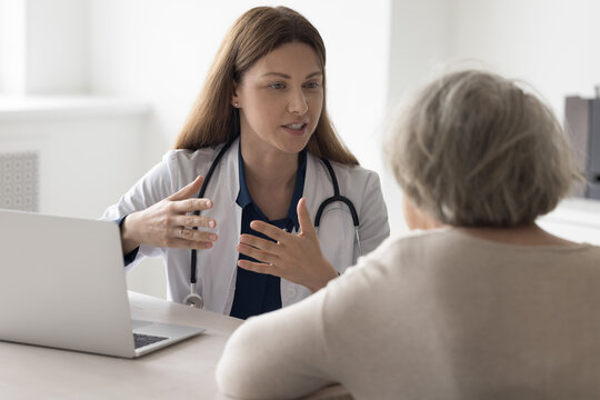 Positive geriatrician doctor woman talking to elderly patient at office workplace, speaking, smiling with hands gestures, meeting with old lady at table with laptop, giving medical help