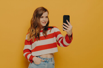 Naklejka premium Young charming woman blogger talking selfie on mobile phone while posing on yellow background