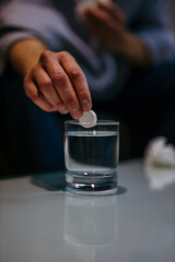Close-up of the man sitting on the sofa in front of the table and throwing the pill into the glass of water he has a headache. Aspirin paracetamol pill splashing into a glass of water.