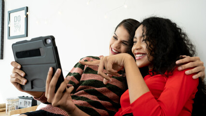 Happy young girlfriends couple hugging having fun using digital tablet relaxing on bed at home. Two smiling women friends holding computer looking at screen enjoying surfing online watching videos.
