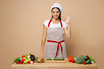 Young happy housewife housekeeper chef cook latin woman wear apron toque chefs hat work at table kitchenware point index finger up isolated on plain pastel light beige background Process cooking food