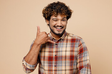 Smiling Indian man wear brown shirt casual clothes doing phone gesture like says call me back wink blink eye isolated on plain pastel light beige background studio portrait. People lifestyle concept.