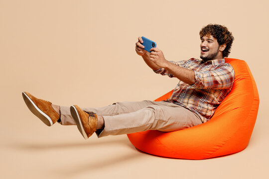 Full body young Indian man in brown shirt casual clothes sit in bag chair use play racing app on mobile cell phone hold gadget smartphone for pc video games isolated on plain pastel beige background - Powered by Adobe