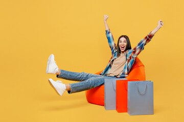 Full body young fun woman wears casual clothes sit in bag chair near paper package bags after shopping do winner gesture isolated on plain yellow background studio. Black Friday sale buy day concept.