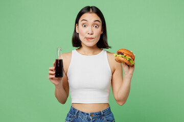 Young shocked fun woman wear white clothes hold bottle of soda pop cola water eat burger isolated...