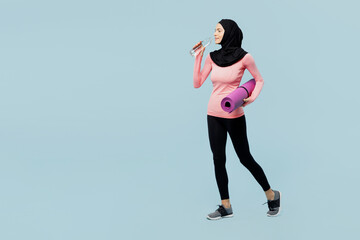 Fototapeta na wymiar Full body young muslim fitness trainer sporty woman wearing pink abaya hijab spend time in home gym walk hold yoga mat drink water walk isolated on plain blue background studio Sport fit abs concept.