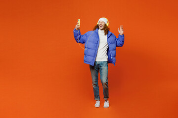 Full body young happy man with long curly hair wearing hat purple ski padded jacket casual clothes headphones listen to music use mobile phone isolated on plain orange red background studio portrait. © ViDi Studio