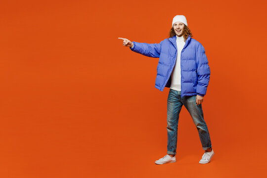 Full body smiling happy cheerful young man with long curly hair wear hat purple ski padded jacket casual clothes walking going point index finger aside isolated on plain orange red background studio.
