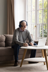 Cheerful older retired man wearing wireless headphones, talking on video call on laptop, writing...