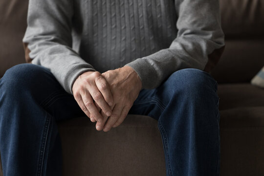 Senior Older Man In Casual Clothes Sitting On Home Sofa, Holding Hands, Folding Palms, Leaning Elbows On Hips, Keeping Pose For Conversation, Body Gesture Of Waiting, Anxiety. Close Up Of Male Hands