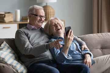 Joyful old husband and wife watching media on mobile phone, talking on video call together, smiling, laughing. Mature retired couple using online app for shopping, internet communication - obrazy, fototapety, plakaty
