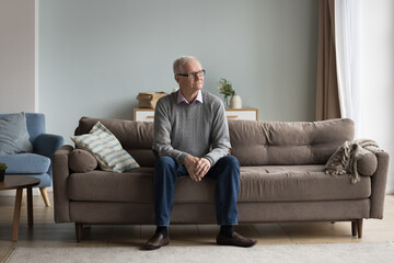 Serious thoughtful old pensioner man sitting on home couch, looking at window away, thinking on health problems, illness, feeling depressed, lonely, anxious. Full length wide shot