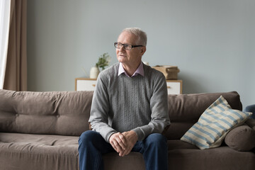 Serious pensive senior pensioner man sitting on home couch, looking at window away, thinking on health problems, feeling sad, lonely, anxious, bored, suffering memory troubles