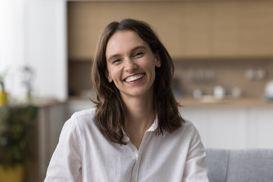 Positive happy pretty young woman home head shot screen portrait. Cheerful adult girl looking at camera with toothy smile, sitting on couch, talking on video call, laughing