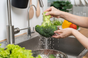 Close up asian young woman washing broccoli, tomato, carrot fresh vegetables, paprika with splash water in basin of water on sink in kitchen, preparing fresh salad, cooking meal. Healthy food people.