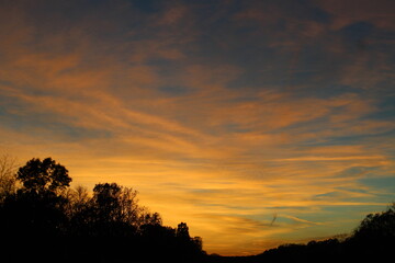 an orange and blue sunset with the silhouette of trees and a bright blue sky and wispy clouds
