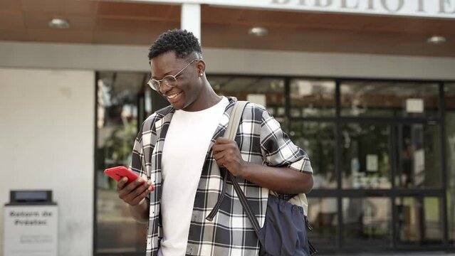Happy African student walking out of University building using cell phone to text friends