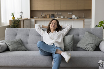 Obraz premium Happy apartment buyer girl relaxing on new comfortable sofa at home, leaning on back with hands on nap, breathing fresh air, enjoying comfort, leisure, looking at camera, smiling, laughing