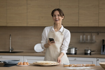 Happy cook blogger girl holding mobile phone over raw bakery food on floury kitchen table, looking at camera, smiling, laughing. Cheerful young baker woman taking picture of food on cell