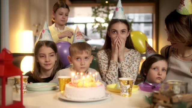 Children's birthday concept. A little boy closes his eyes to make a wish in front of a birthday cake with burning candles. Festive table, birthday paraphernalia. Waiting for a miracle