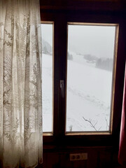 Fenster einer Alphütte, zeigt in eine schöne verschneite Winterlandschaft in den Bergen. Fenster geschlossen..Weiße retro Gardine verdeckt die Sicht an  einer Seite.