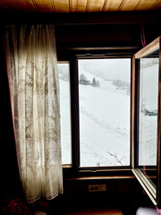 Fenster einer Alphütte, zeigt in eine schöne verschneite Winterlandschaft in den Bergen. Fenster auf einer Seite geöffnet.Gardine hängt an der geschlossenen Seite