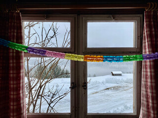 Fenster einer Alphütte, Zeigt in eine schöne verschneite Winterlandschaft in den Bergen. Fenster ist verschlossen. Karierte Vorhänge, rechts und links, Regenbogen farbige  Girlande.Holzdecke