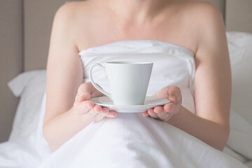 Woman in bed holds a white cup on a saucer in her hands. Bare-shouldered woman got breakfast in bed. The concept of taking care of yourself, vacation, have a nice morning.