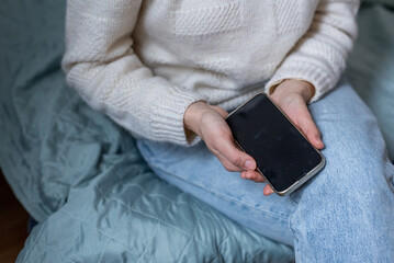 Young woman in white sweater and blue jeans sitting on the blue sofa and holding in her hands her mobile phone.