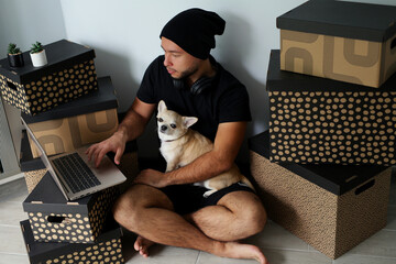 A young, stylish guy in black clothes is at home with his dog. A Chihuahua dog and a guy watch a movie together on a laptop, have fun and spend time together, real friends. The guy and the dog are in 
