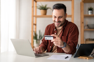 Young businessman millennial man consumer working on laptop holding credit card sitting at desk at...