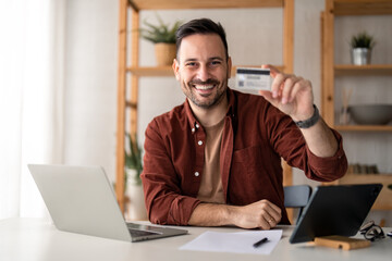 Smiling businessman holding and showing credit card while sitting in home office. Male business manager advertising online internet banking services, loan or deposits, cashback for purchases concept.