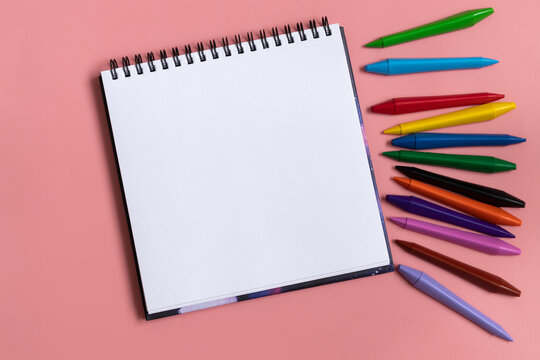 Open sketchbook with white empty blank. Colorful wax crayons on the pastel pink background. Flat lay composition.