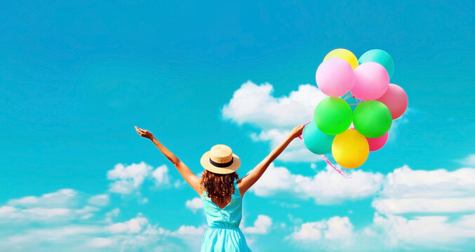 Back view of happy smiling young woman with bunch of colorful balloons wearing summer straw hat on blue sky clouds background