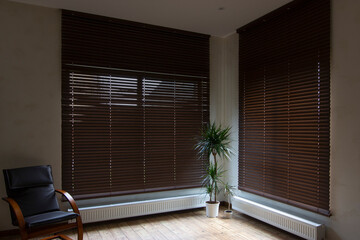 Wooden blinds on large windows in the interior. Living room with armchair and houseplants near...