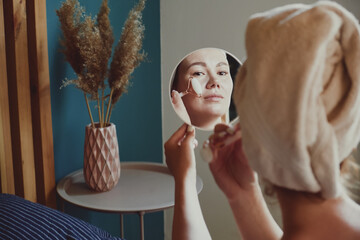 Close up mirror, woman using jade facial roller for face massage sitting on bed in bedroom looking...