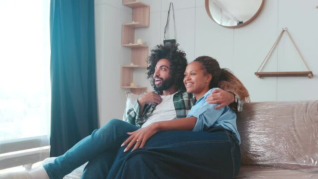 Young dreamy Arabian man and African American woman hugging with smile imagining happy future after relocation to new house with panoramic windows sits on sofa in lounge with boxes. Multiethnic family