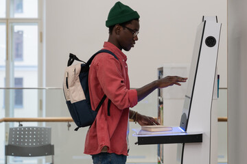 Young African student guy with backpack standing in library using self-service terminal to borrow...
