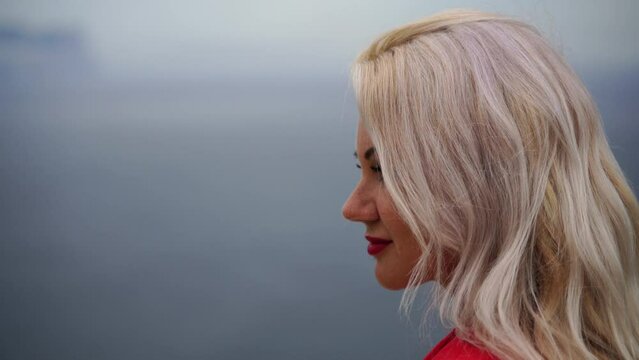 Portrait blonde sea beach. A woman is sitting by the sea in a red jacket and red lipstick, enjoying the sea view.