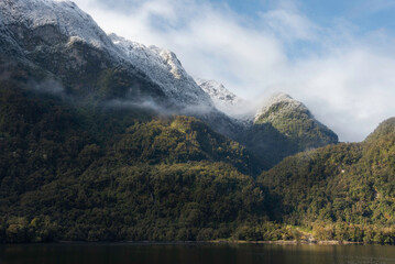 Fototapeta na wymiar The diverse shores in the Doubtful Sound with spotlights of light, wispy clouds, and snowcapped rainforest.
