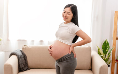 Young asian beautiful pregnant woman holds hands on belly standing near window at home. Pregnancy, maternity, preparation and expectation concept.