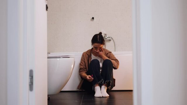 Anxious young white woman with nervous feelings on bathroom. Caucasian girl holding a positive pregnancy test, crying after finding result. Pregnancy test kit in hands. Emotional female in restroom.